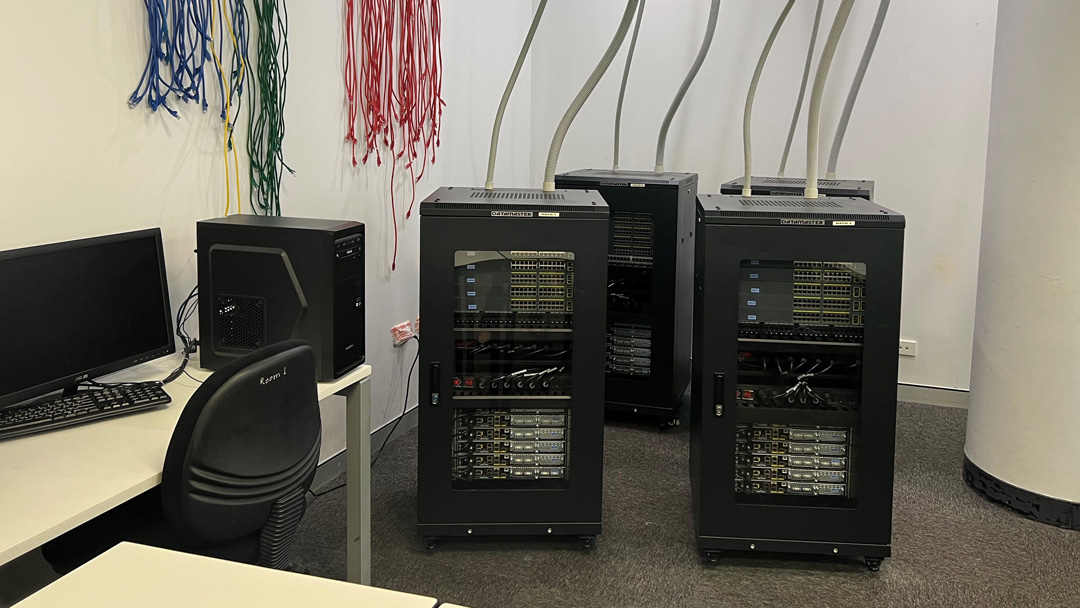 Four servers with pipes of wires flowing into the ceiling. Next to the servers are a computer sitting on a desk.