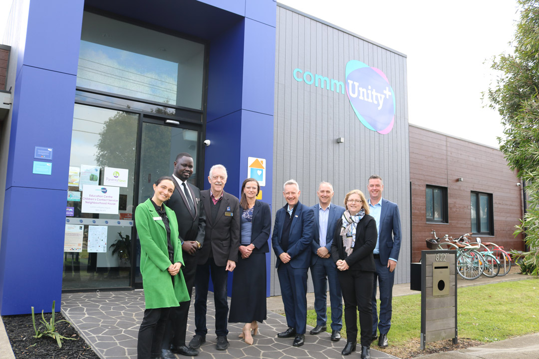 A group of people standing outside, looking at the camera. A building with the commUnity+ logo on it.