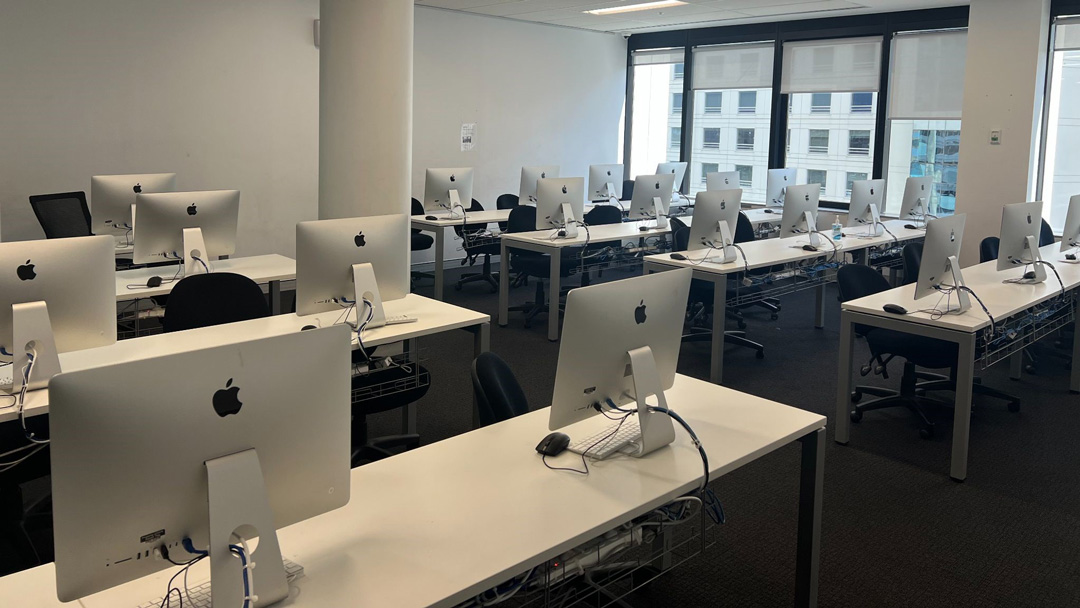 Four rows of desks split down the middle. Each desk has a Mac computer. To the right are tall windows that view another building.