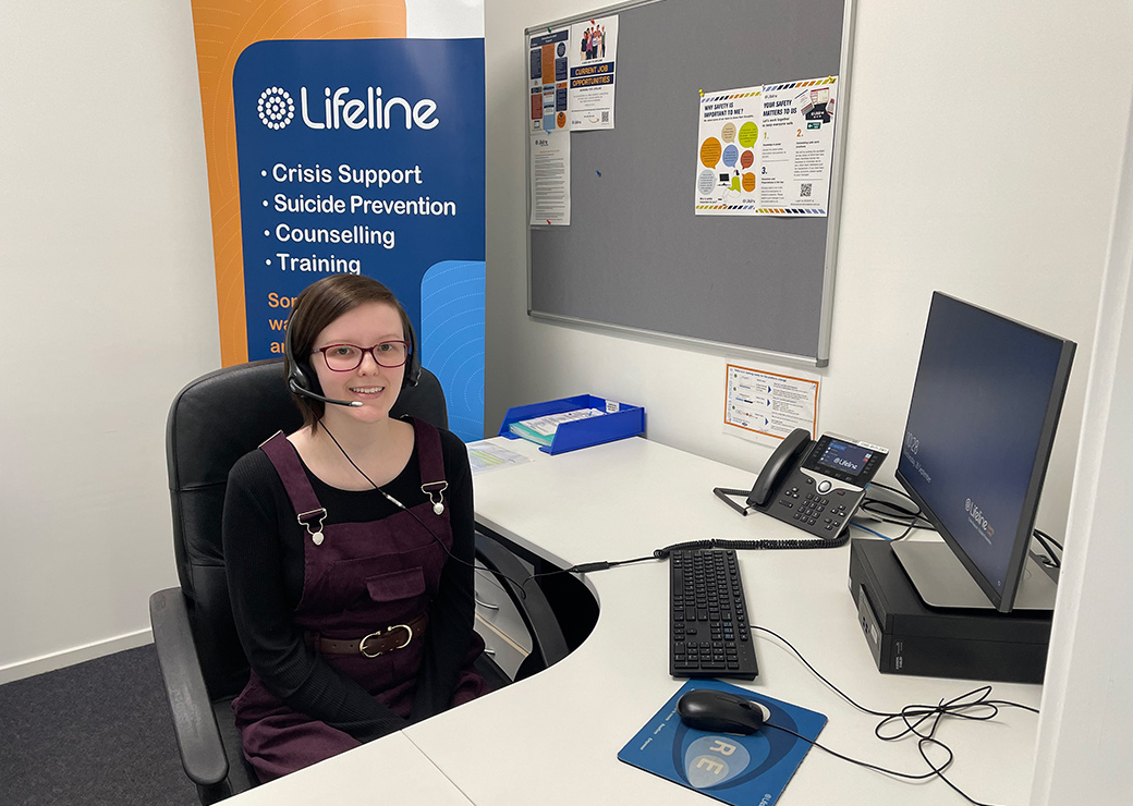 Young woman in casual clothing, wears a microphone headset at an office desk with Lifelife poster 