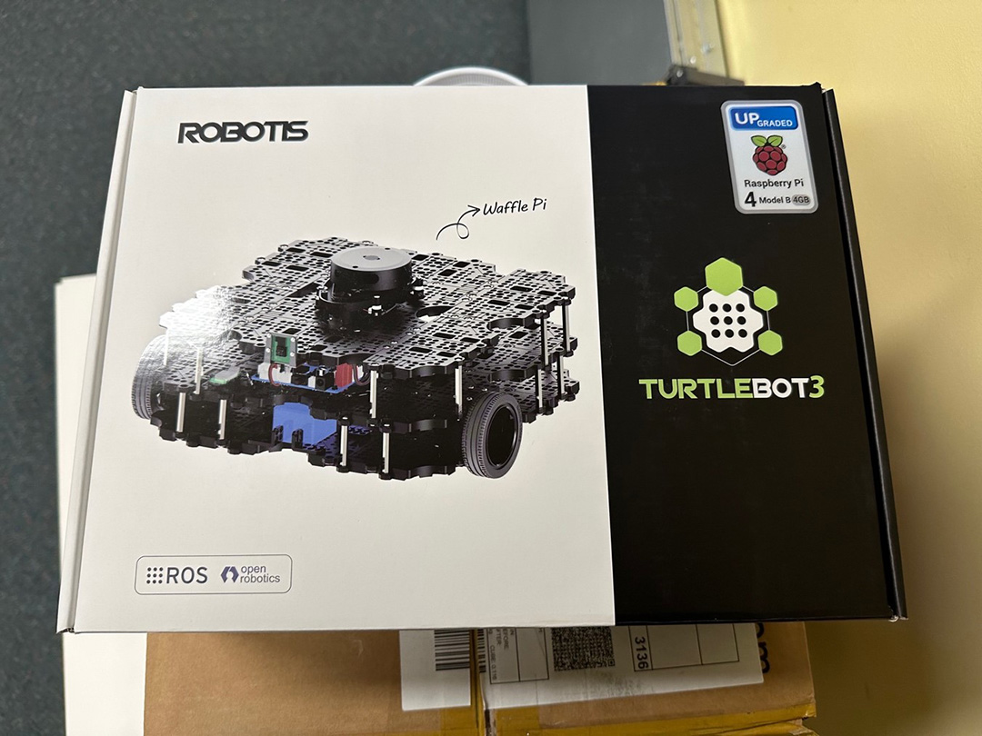  A closed cardboard box. The box has a picture of a small square robot with wheels. The box says "Robotis" and "Turtlebot3" with a Raspberry Pi logo.