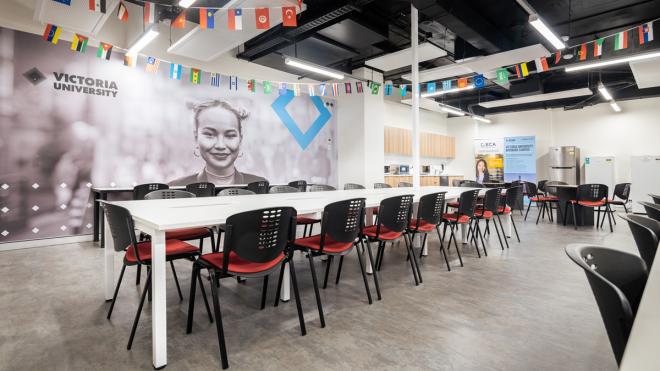 Large room with chairs and a long table. In the background is a fridge and kitchen, with several Victoria University banners surrounding it. Several different countries' flags hang from the ceiling.