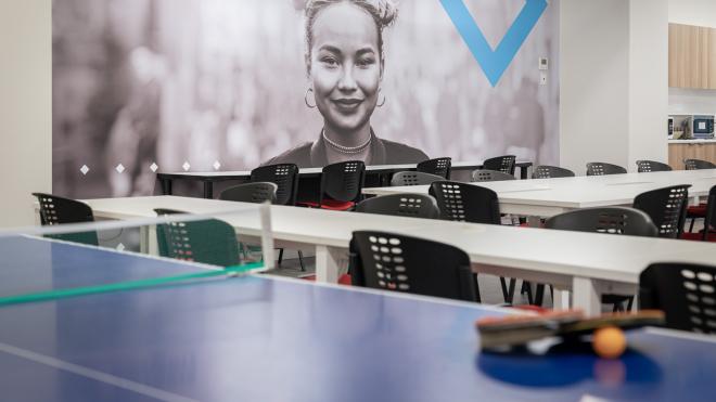  A table tennis racquet and ball sitting on a table tennis table. In the background are tables and chairs.