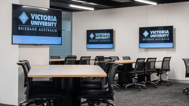  A large room with long tables and computer chairs surrounding them. Each table has a computer screen next to it that is mounted to the wall.