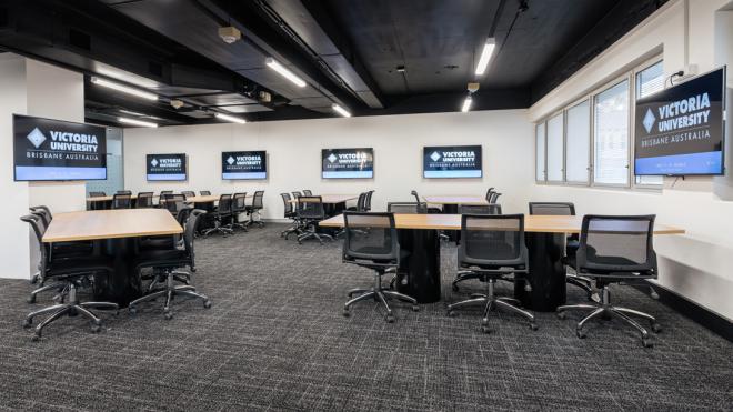  A large room with long tables and computer chairs surrounding them. Each table has a computer screen next to it that is mounted to the wall.