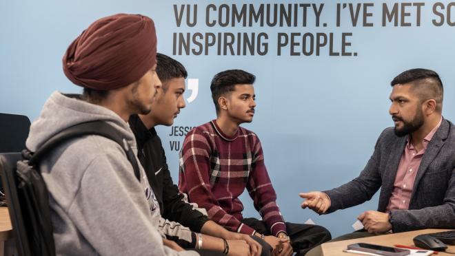 Four people sitting down. One person is talking to them, using their hands to make gestures, while the other three listen to them.