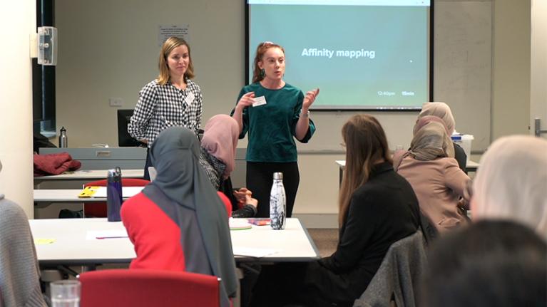 Two presenters speaking to a powerpoint in front of several students sitting at desks