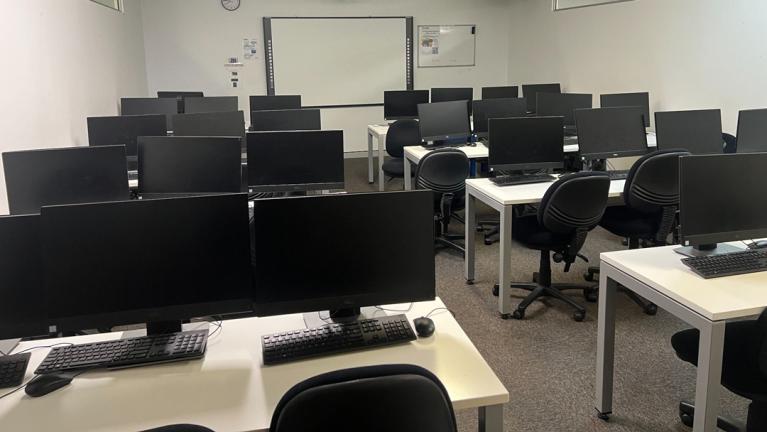  Four desks split down the middle. Each desk has computer monitors, keyboards and mice on them. At the back of the room is a digital whiteboard.