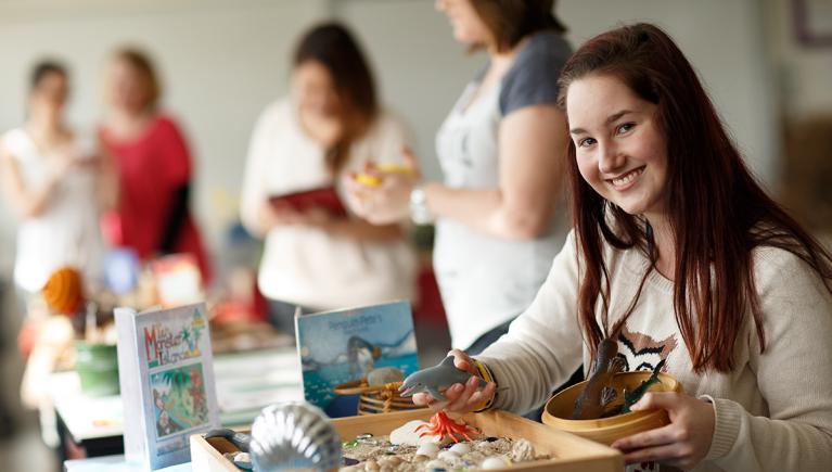  Education student smiles to camera as she sits at table with craft materials, whilst other students mill in the background