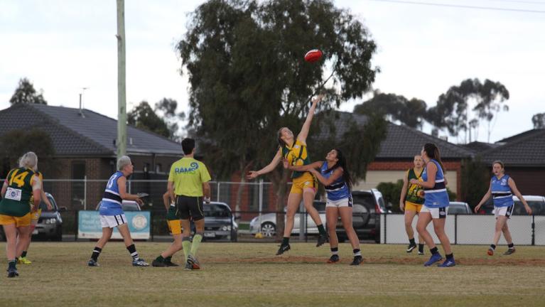 Football players on a field - houses in background. A player is jumping and reaching for the ball in the air, another player is next to them looking at the ball.