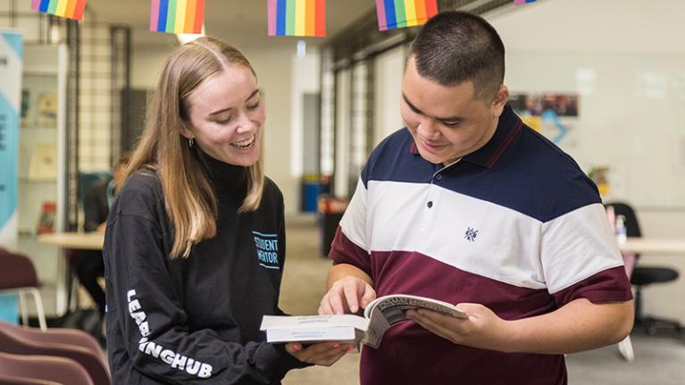 Female student in mentor shirt, smiles and checks reference book with male student