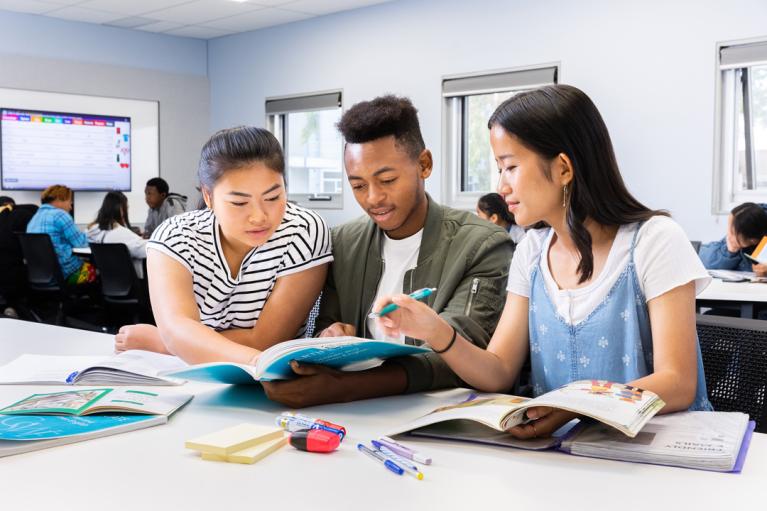  Students in a classroom. Three students in the foreground are sitting down and looking at the same book.