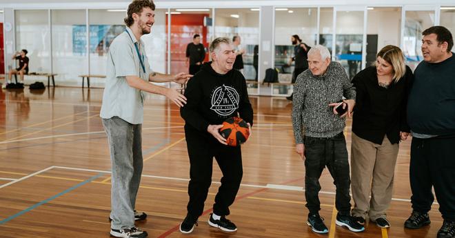 A man holding a basketball preparing to shoot. A man stands behind him, holding him. Three people are watching by the man's side.