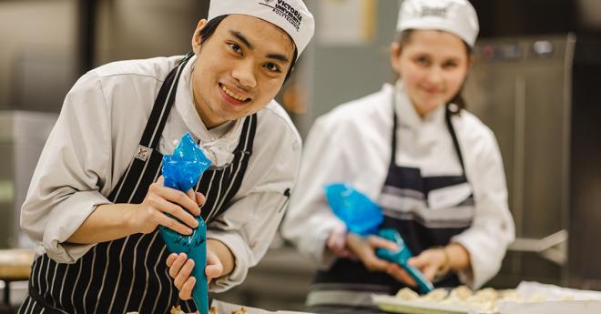 A young man and woman in chef uniforms smile while adding sauce to a platter