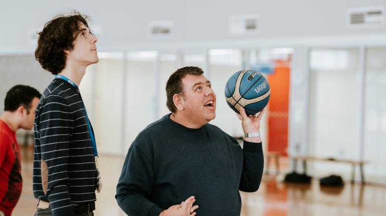  A person holding a basketball in their left hand, looking up and positioning themselves to shoot. A person stands to their right, looking in the same direction.