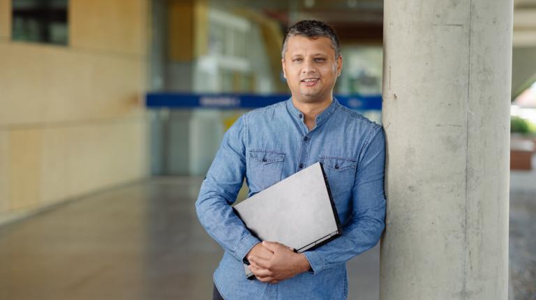  student leaning against a column, relaxed, holding laptop