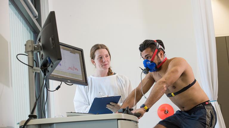 man hooked up to monitoring equipment rides an exericise bike, watched by a woman in a lab coat