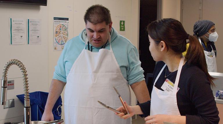  Male participant in CHEW program with VU nutrition student holding a pair of tongs explaining preparation of a wrap.