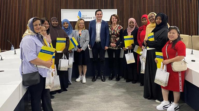 A group of women in variety of cultural dress hold certificates with a Moonee Ponds logo in the background