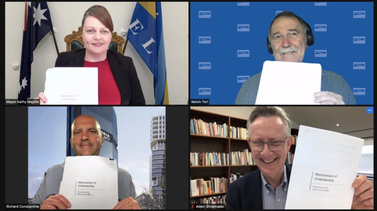 Zoom meeting with 4 professionals holding up an official agreement, with flags, logos and bookshelves in the background