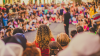  Crowds standing and sitting in a circle around a figure, blurred in the background.