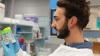 A young male scientist in a white lab coat examines 2 sample containers