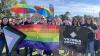 Group of men and women holding rainbow umbrellas, flag with Victoria University logo, at an outdoor gathering